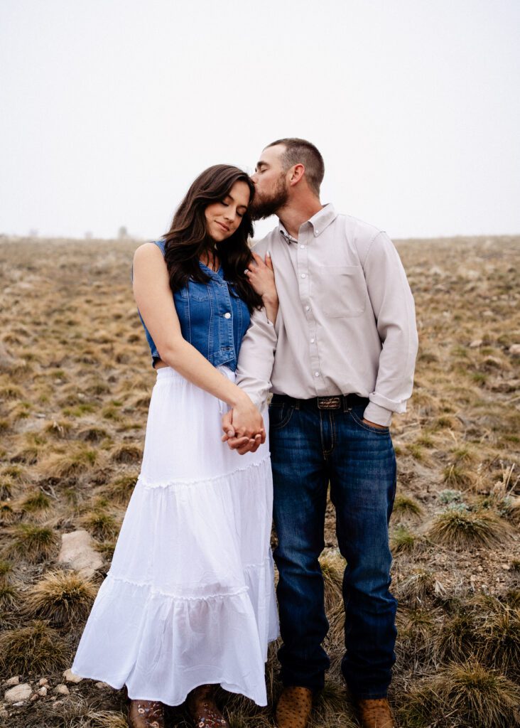 fiance kissing his girl friend on the head as they hold hands and close their eyes in a field during their colorado proposal photoshoot