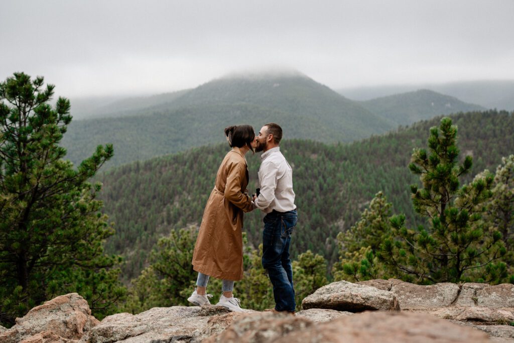 Girl reaching up to her boyfriend after he just proposed to her on a mountain in colorado with a foggy mountain landscape in the background