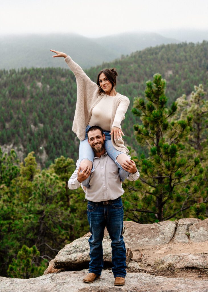 girl sitting on her fiances shoulders as she shows off her engagement ring with the mountains in the background during their proposal in colorado