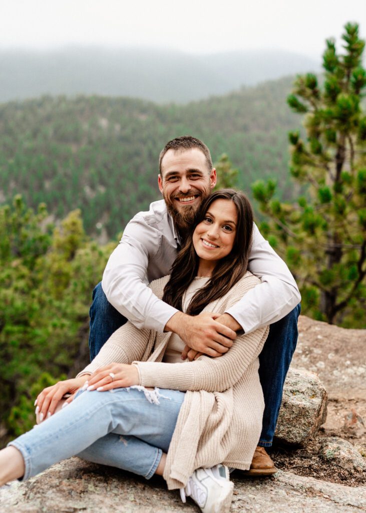 fiance hugging his girl friend from behind while smiling at the camera with the mountain in the background during their proposal in colorado