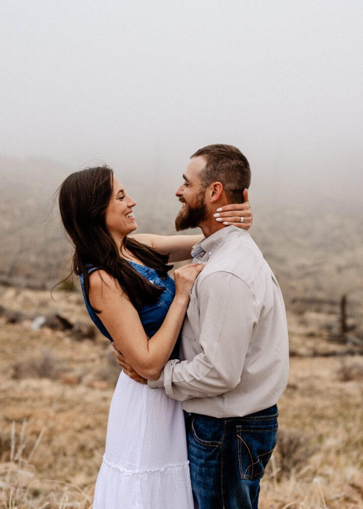 newly engaged couple standing in a field smiling at each other during their proposal session in colorado