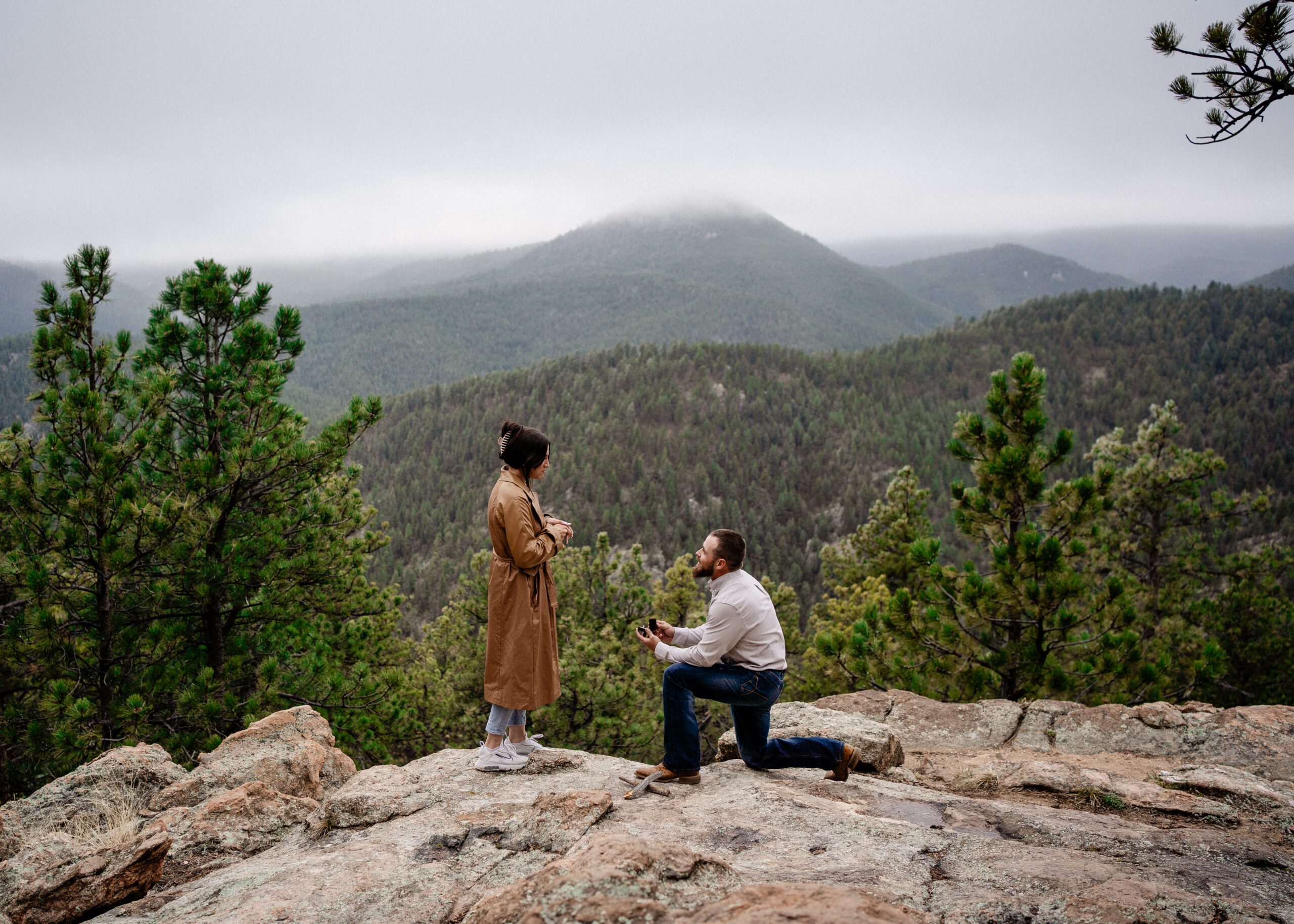 Man down on one knee proposing to his girlfriend on top of a mountain during their proposal in colorado