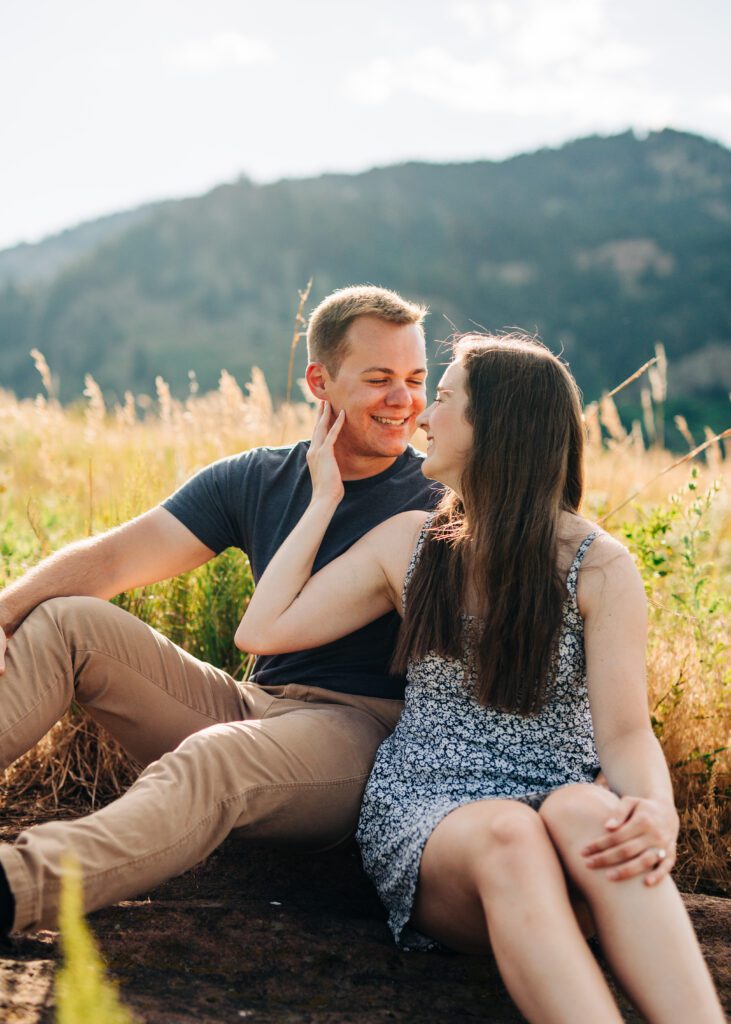 girlfriend holding her boyfriends face as they smile at each other while sitting in a field during sunset during their boulder engagement session