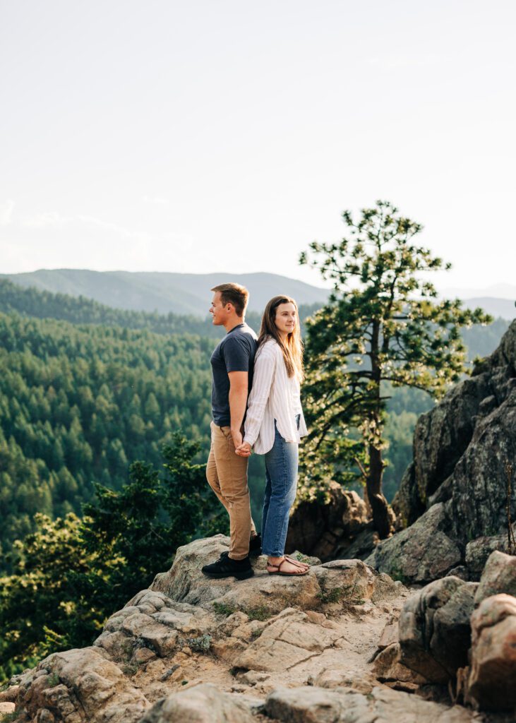 engaged couple standing back to back on a rocky cliff at lost gulch overlook in boulder colorado during their couples photoshoot