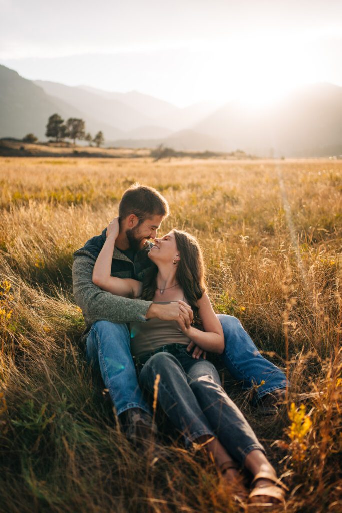 cute engaged couple sitting in a field together while smiling and laughing during their engagement photos in rocky mountain national park