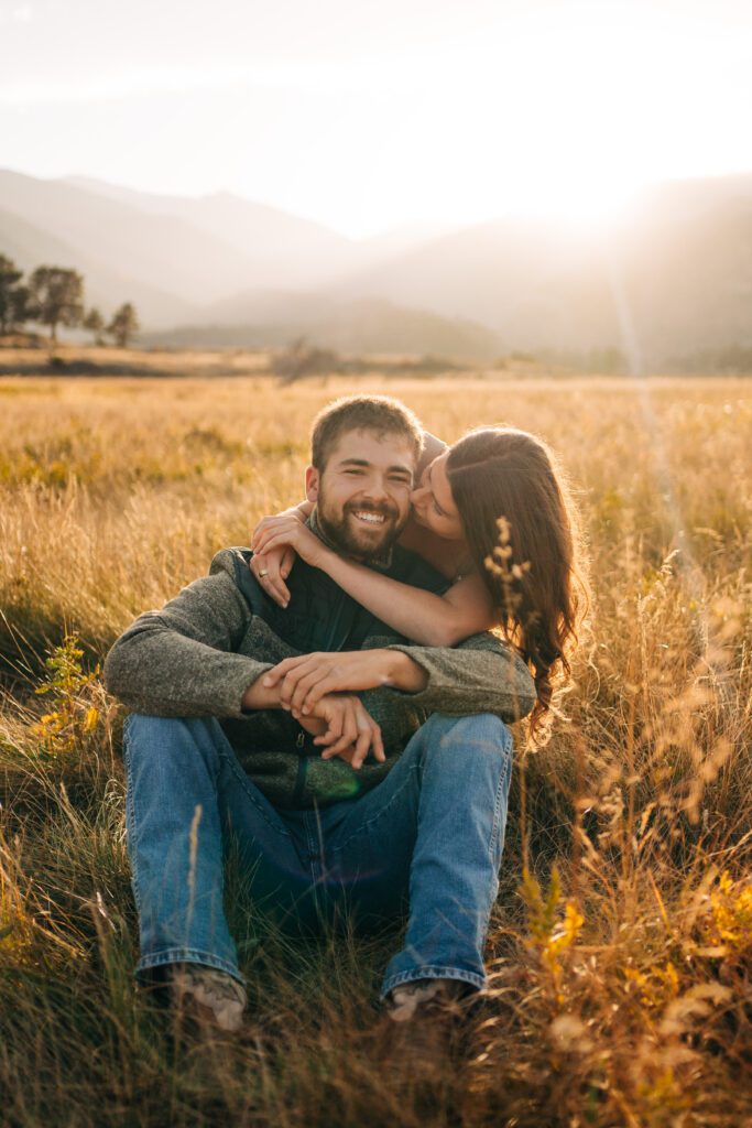 corwin smiling at the camera while kadi kisses him on the cheek during their engagement session in rocky mountain national park