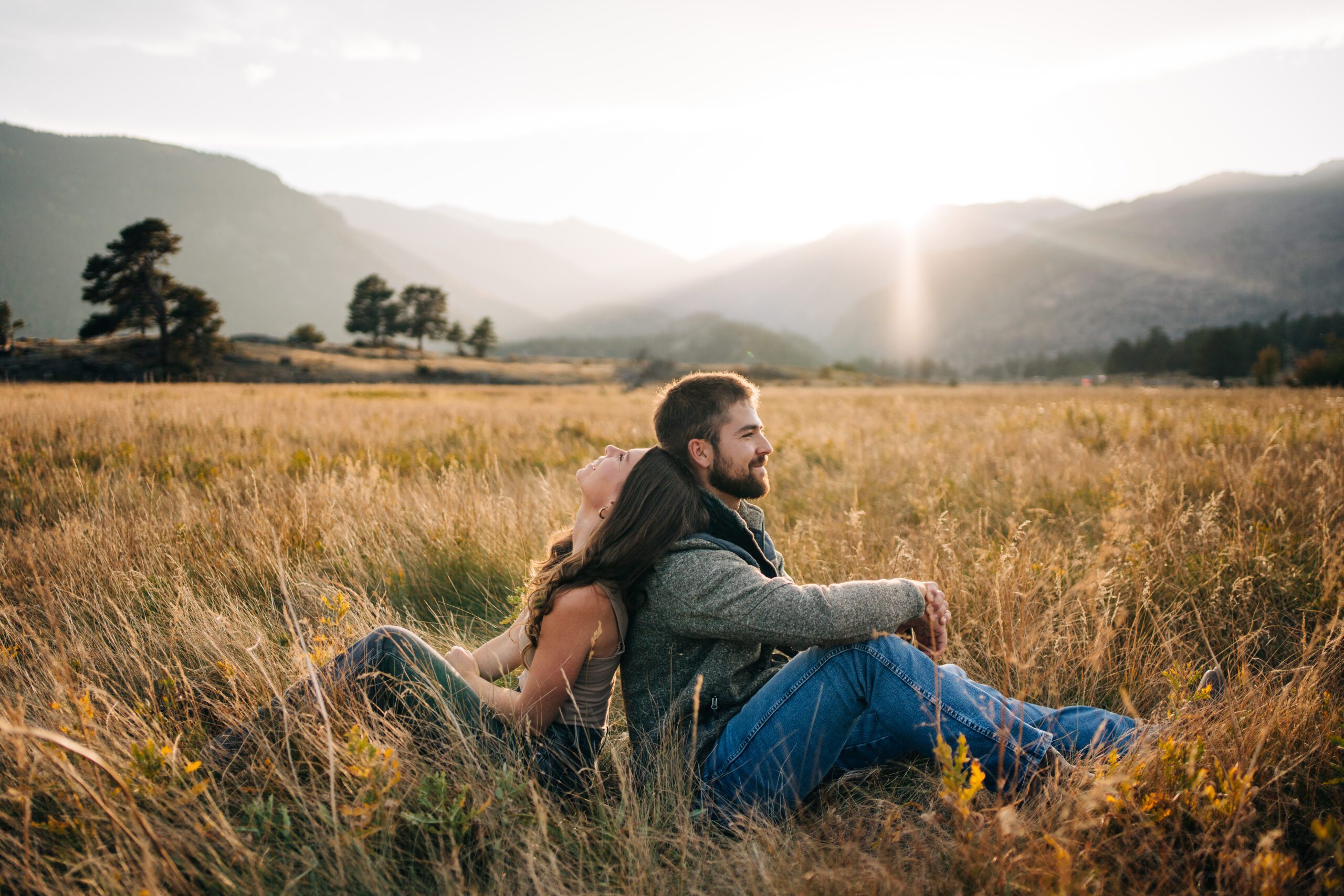 Cute engaged couple sitting back to back in a field in Moraine Park during their engagement session in rocky Mountain National park