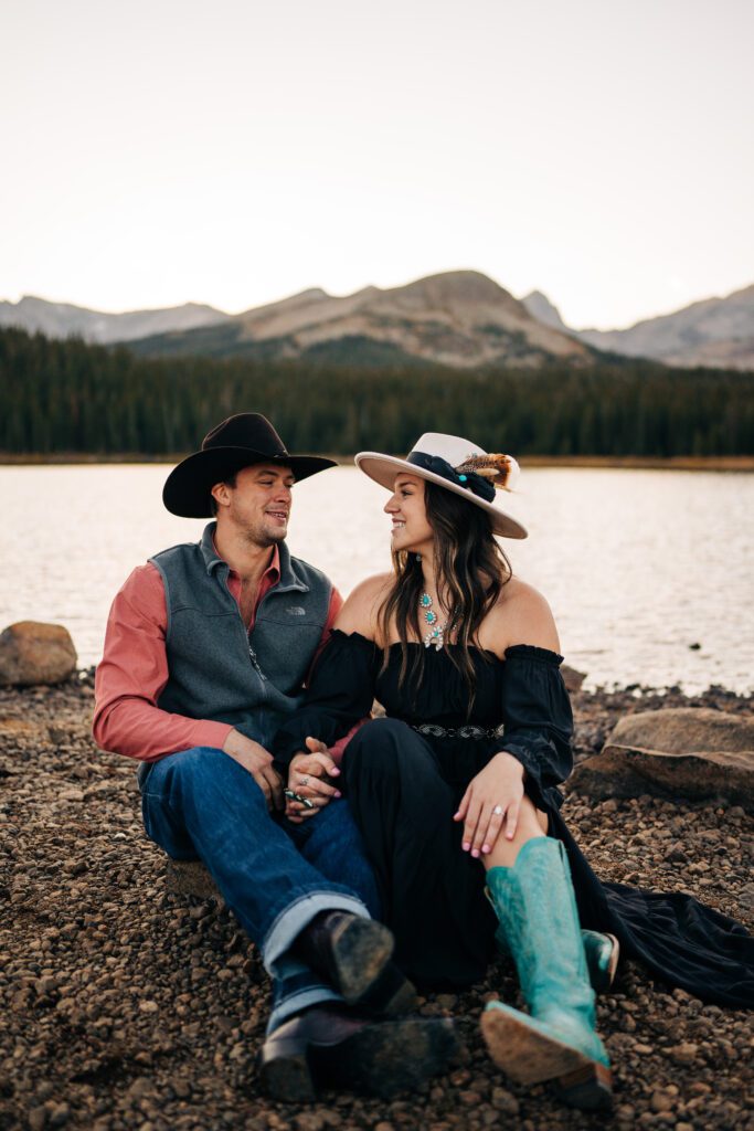 Engaged couple sitting on the lakeshore smiling at each other during their sunset Brainard Lake Engagement Session in Colorado