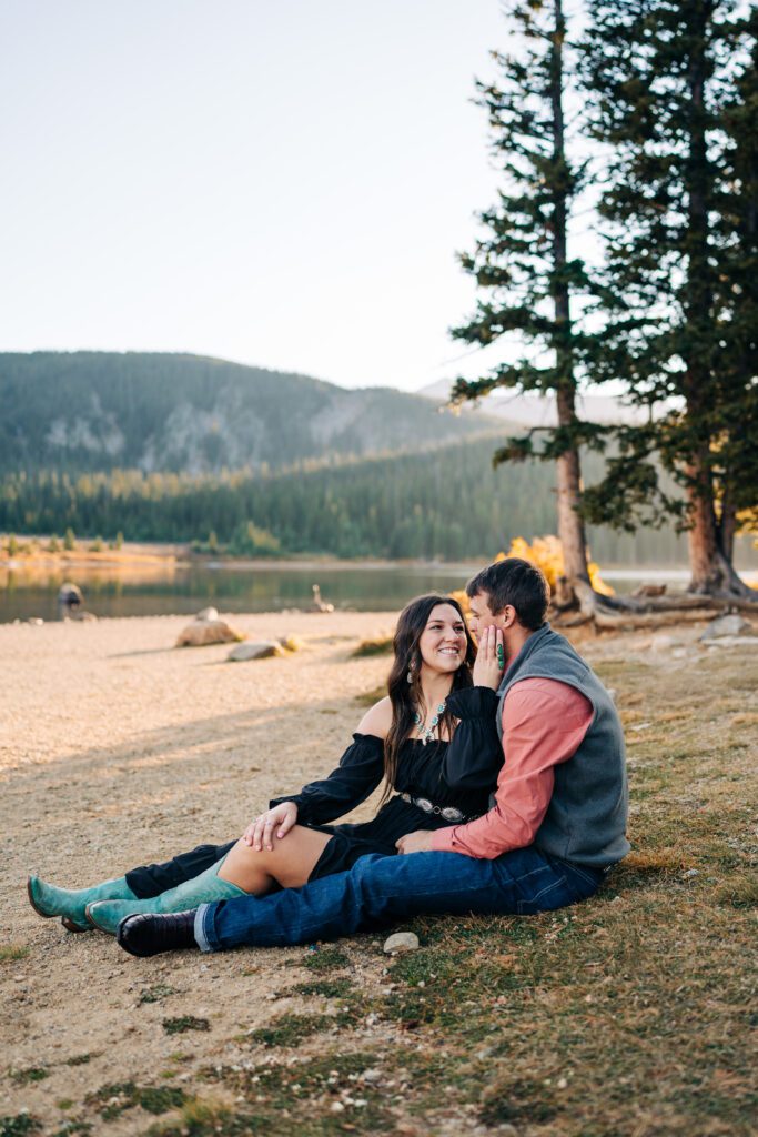 Cute engaged couple sitting together on the shoreline of a lake during their Brainard Lake engagement session in Colorado