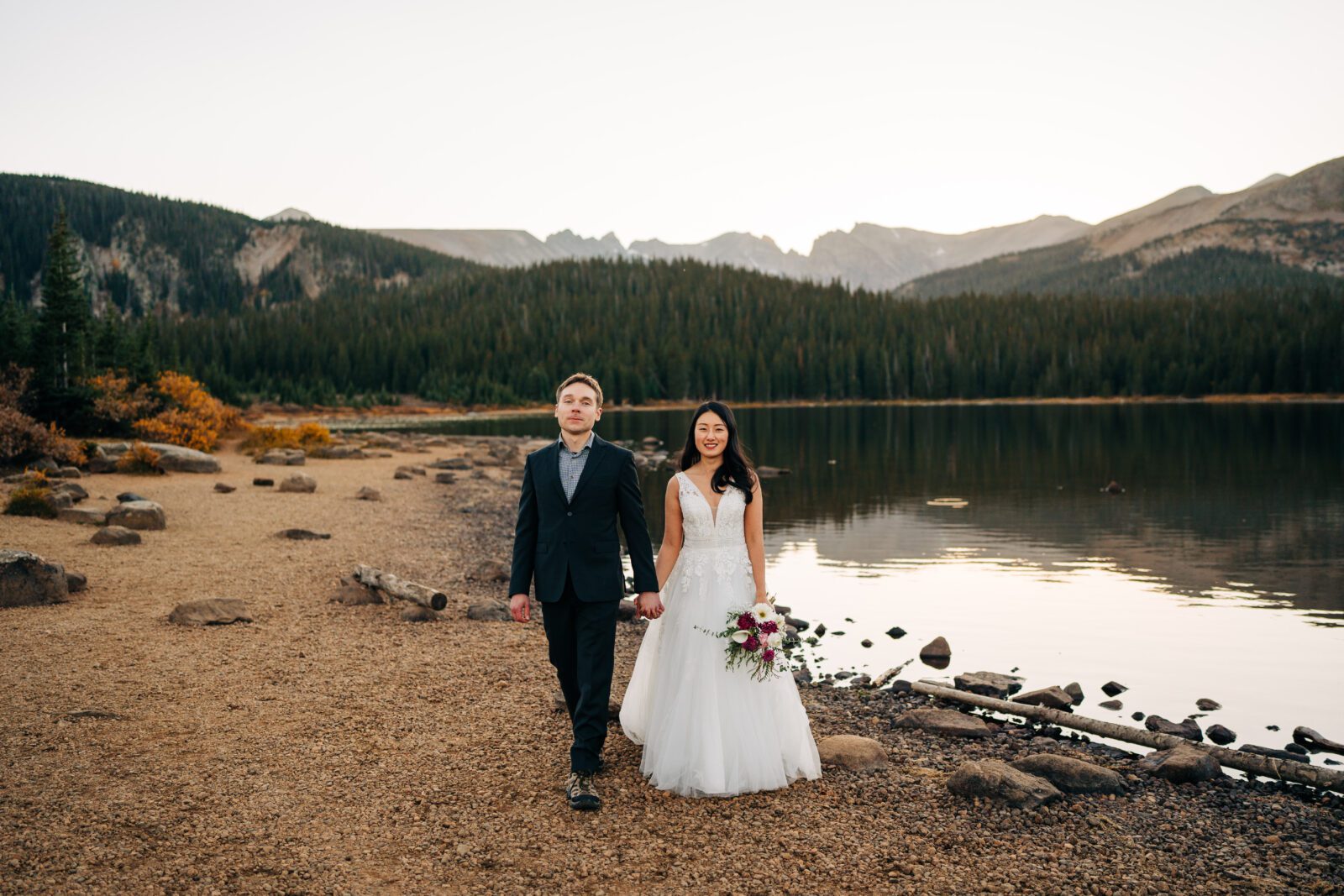 bride and groom holding hands and walking towards the camera along the lakeshore during their sunset brainard lake elopement near boulder colorado