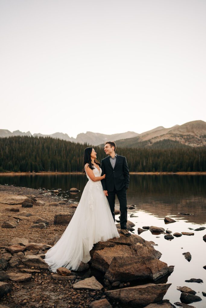 bride grabbing grooms arm while they smile at eachother while standing on a rock with the mountains and lake in the background during their brainard lake elopement near boulder colorado