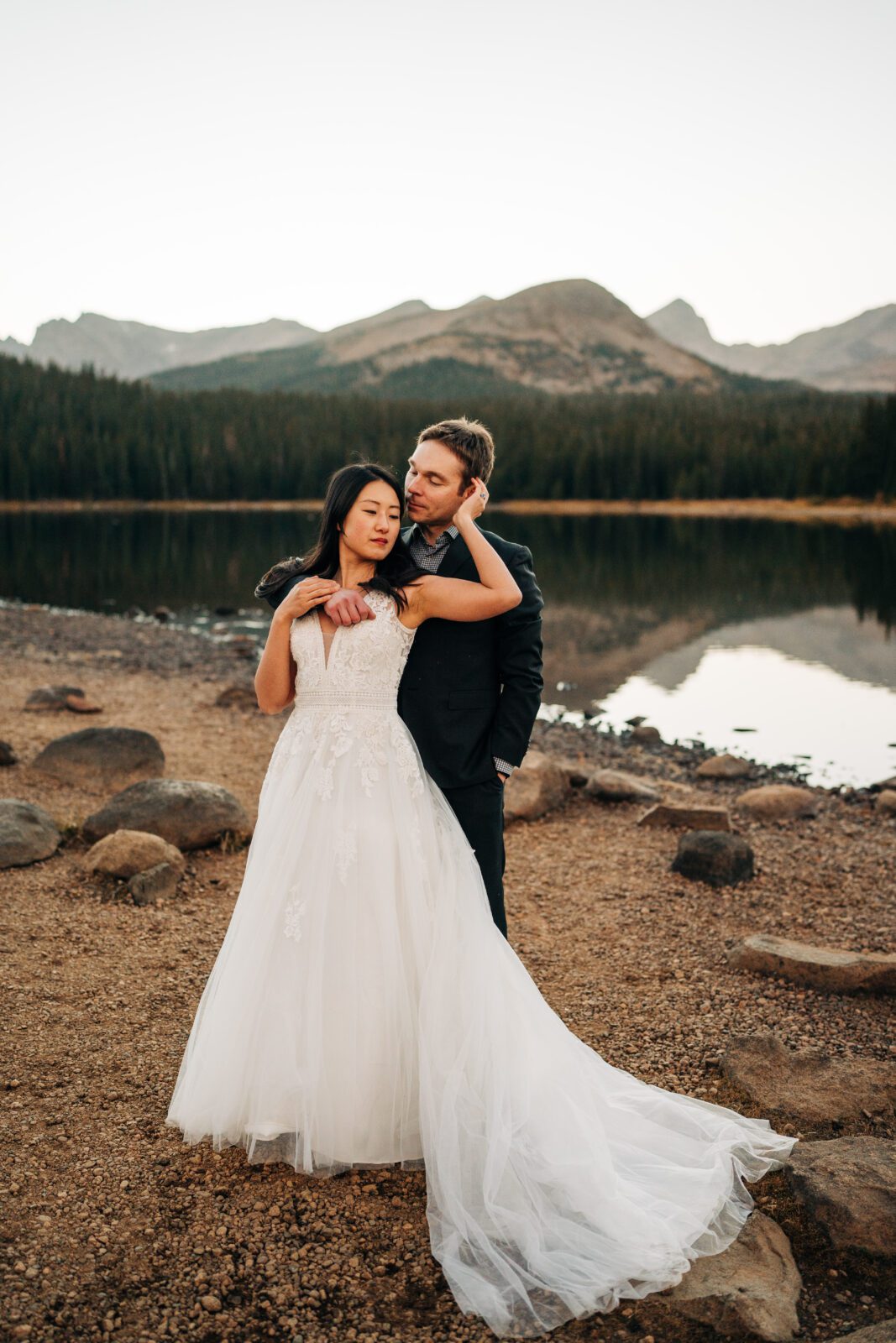 bride standing in front of the groom laying back on him while reaching up to his face and looking off in the distance with the mountains in the background during their brainard lake elopement near boulder colorado