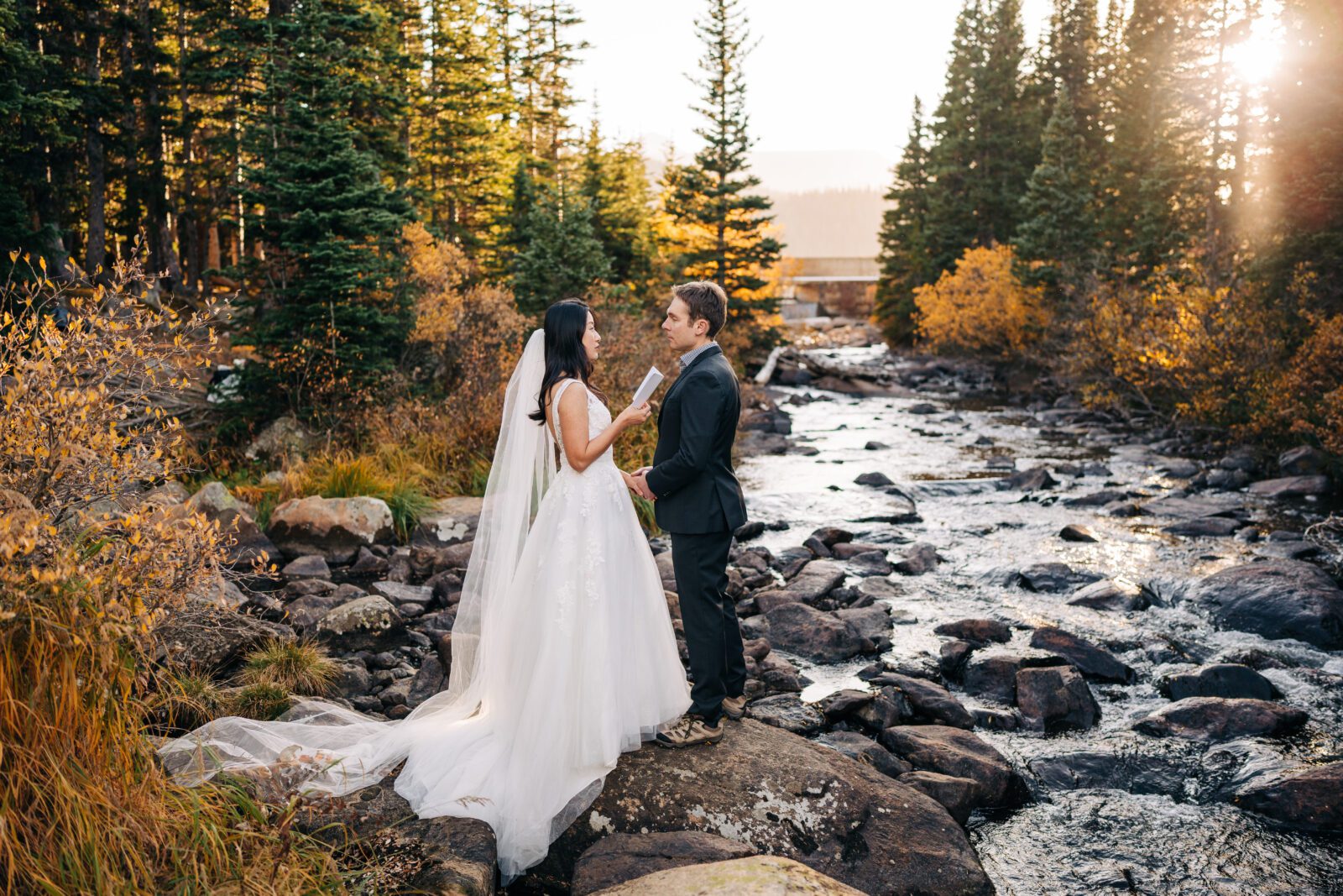 bride and groom standing in the forest next to a river at sunset while the bride reads her vows to the groom during their brainard lake elopement 