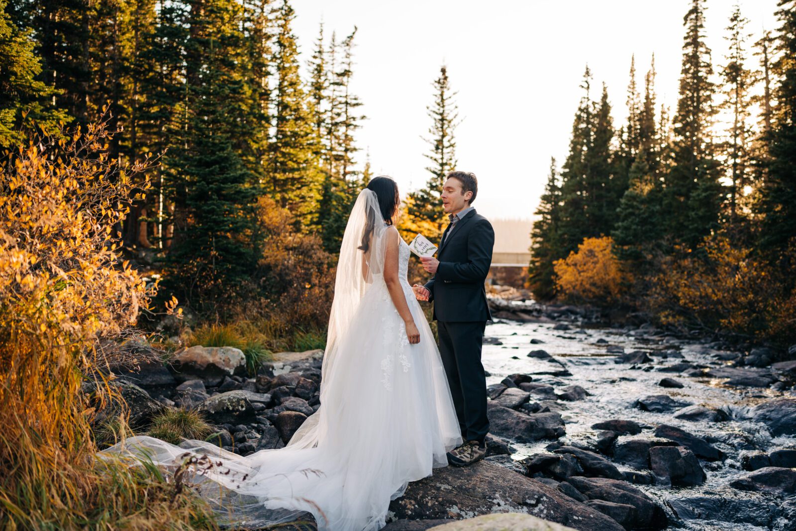 groom reading his wedding vows to his new wife during their brainard lake elopement in the forest at sunset with a river next to them