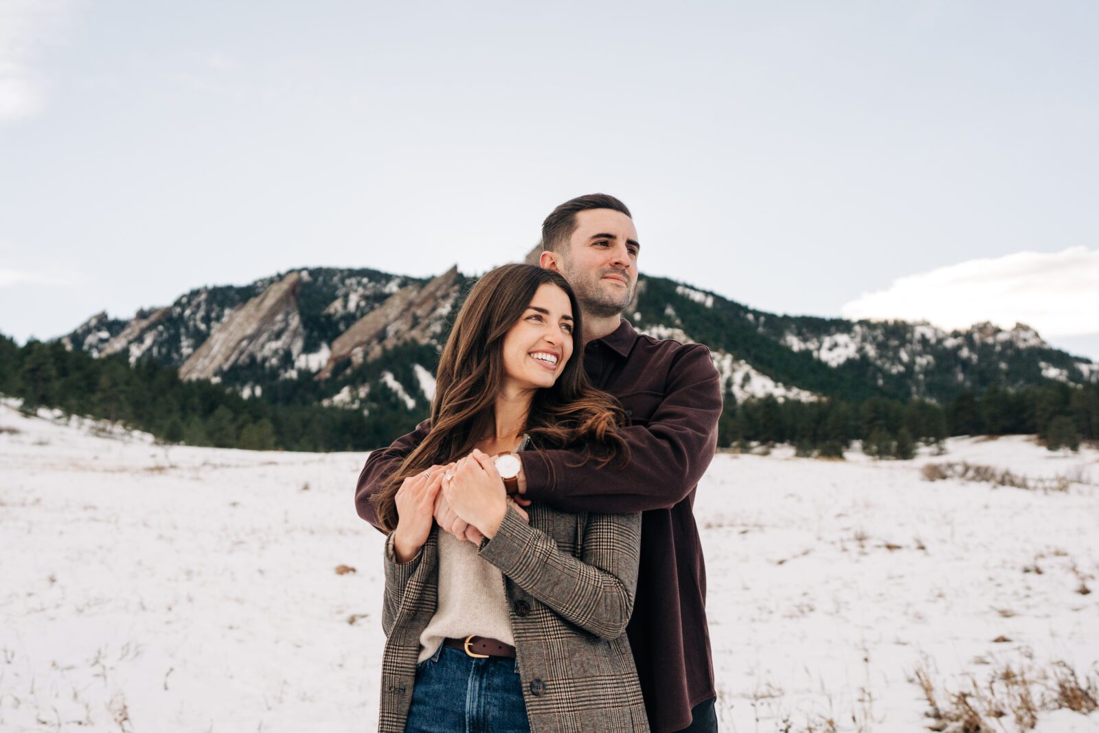 Newly engaged couple holding eachother and looking off in the distance with the Boulder Flatirons behind them during their Colorado winter engagement photoshoot