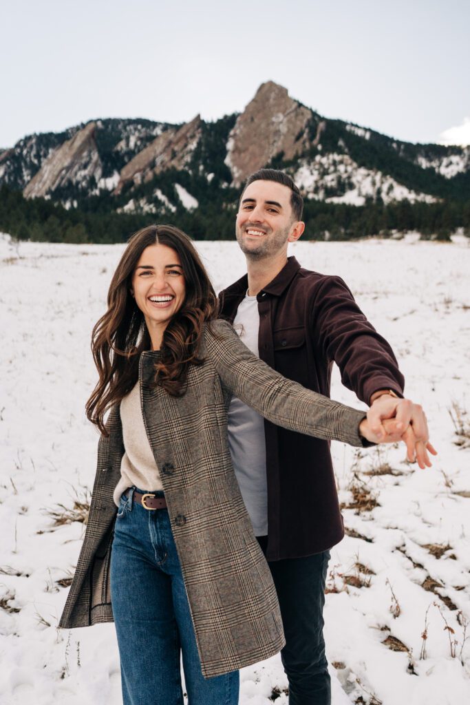 Fiance holding his girlfriends hands and swinging them in an airplane motion in a snowy field at the Boulder Flatirons during their Colorado winter engagement photoshoot