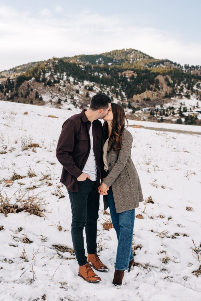 Newly engaged couple holding hands and kissing in a snowy field at Chautauqua Park in Boulder, Colorado during their winter engagement session