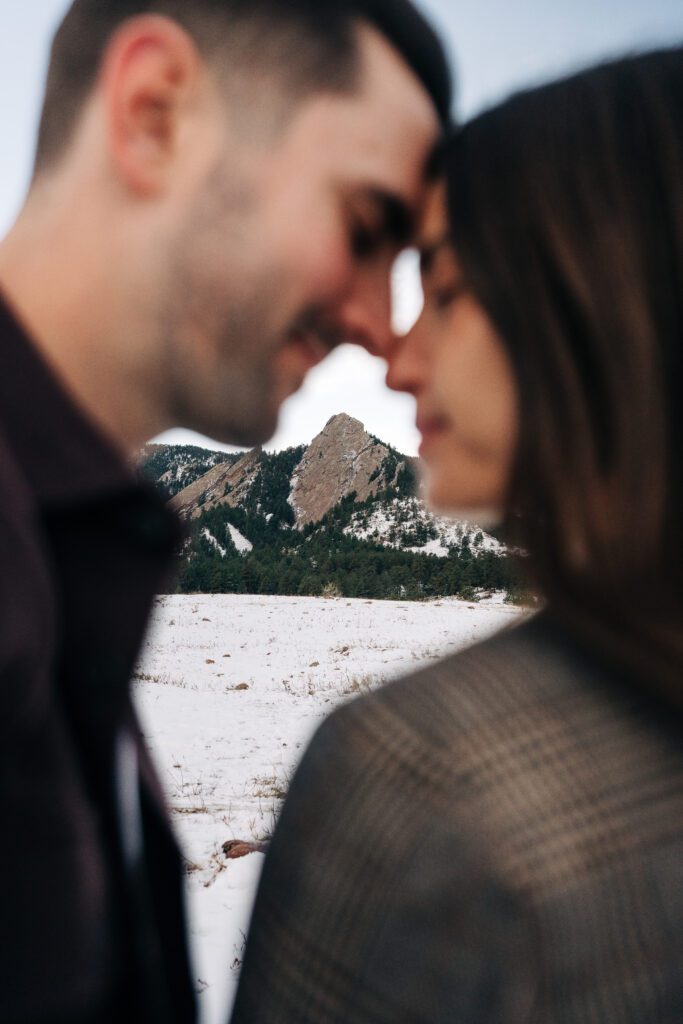 Cute engaged couple with their noses touching and the flatirons focused between their faces at Chautauqua Park in Boulder, Colorado during their winter engagement photos