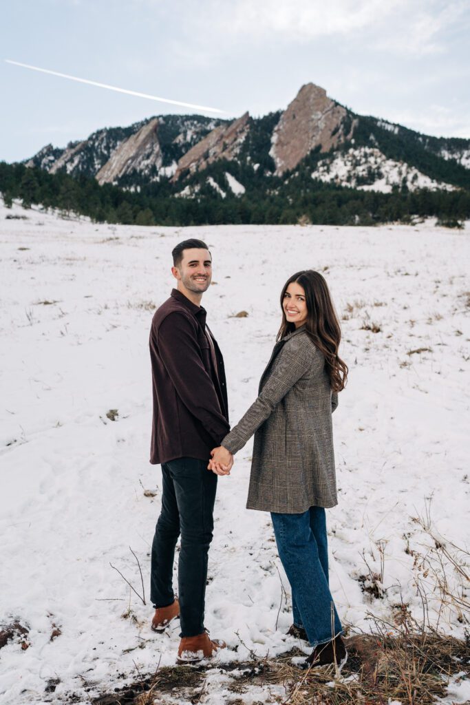 Engaged couple holding hands and walking away from the camera and looking back during their Colorado winter engagement session