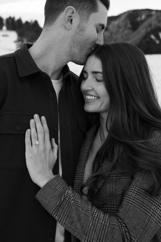 Black and white image of boyfriend kissing his girlfriend on the head with her engagement ring in focus during their winter engagement photoshoot in Boulder, Colorado at Chautauqua Park