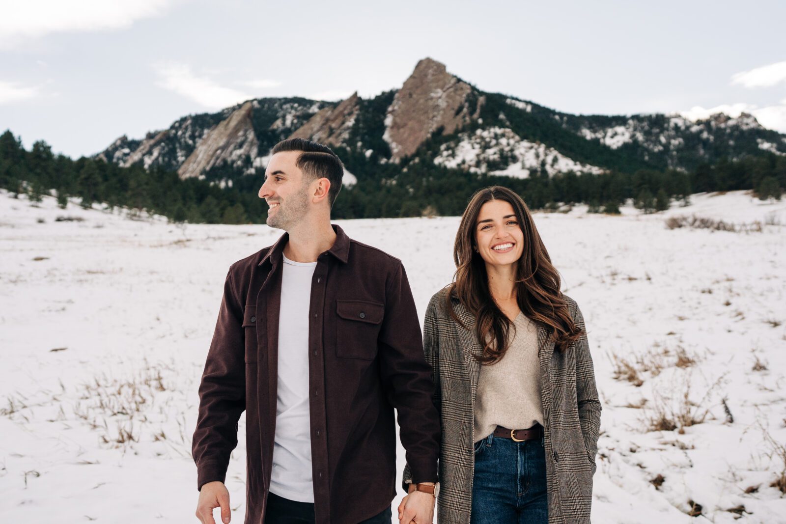 Fiancee smiling at the camera while her fiance looks off in the distance with the Boulder, Colorado Flatirons in the background during their winter engagement session at Chautauqua Park