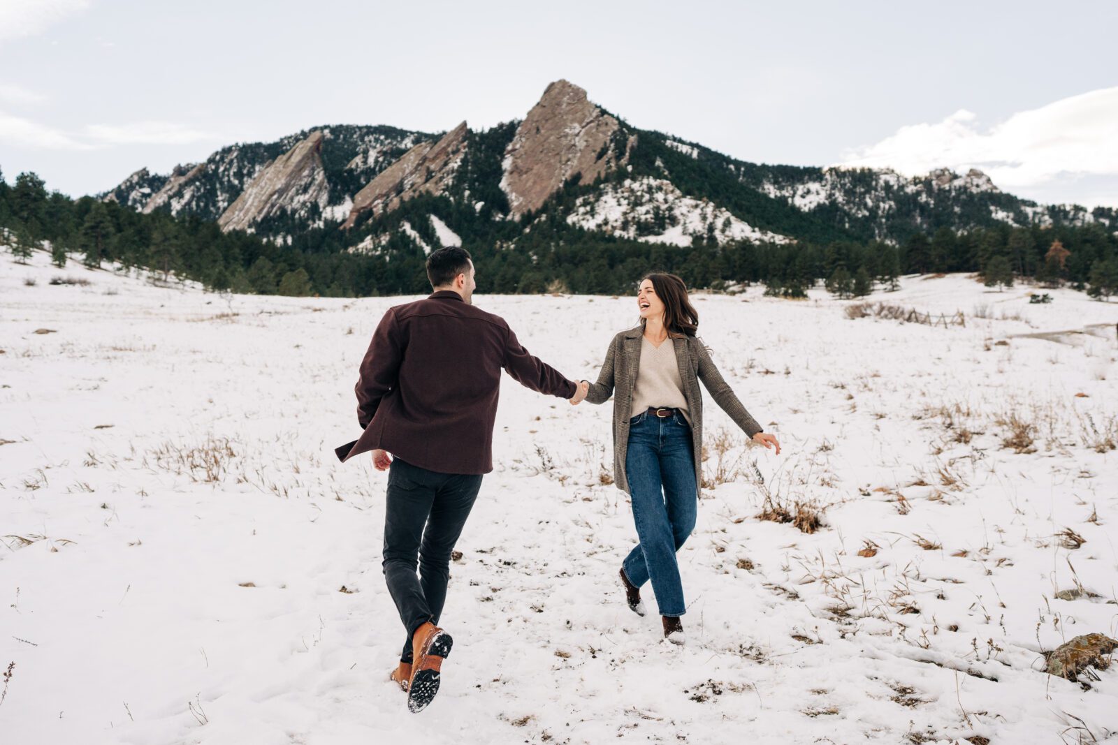 Cute engaged couple holding hands and running around smiling at each other in a snowy field at Chautauqua Park in Boulder, Colorado during their winter engagement photoshoot