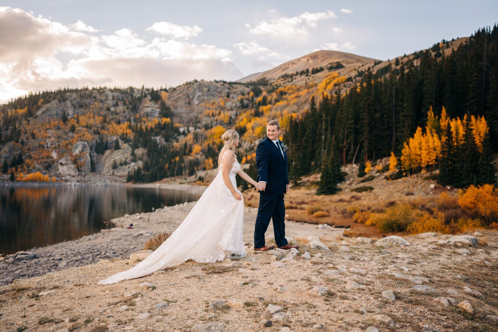 groom holding brides hand as he leads her down a walking path in the Colorado mountains next to an alpine lake with beautiful fall colors in the backdrop during their Breckenridge elopement photos