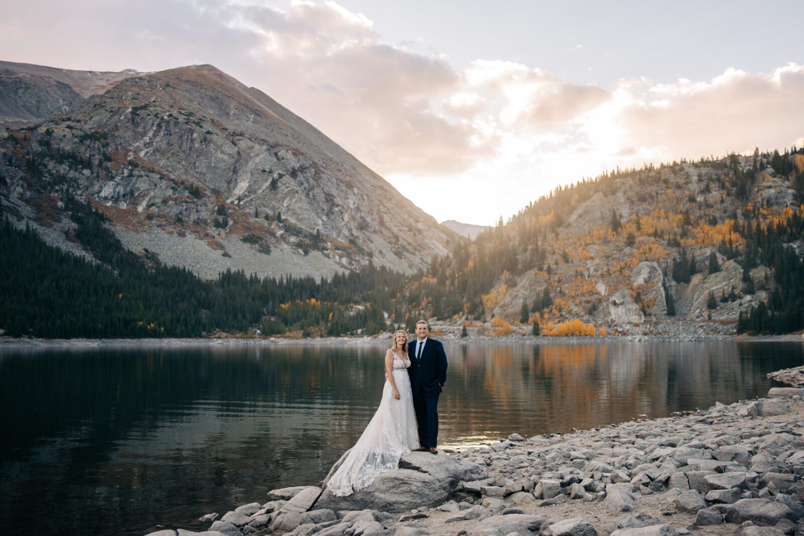 bride and groom standing on a rock on the lakeshore of an alpine lake at sunset during their Breckenridge elopement