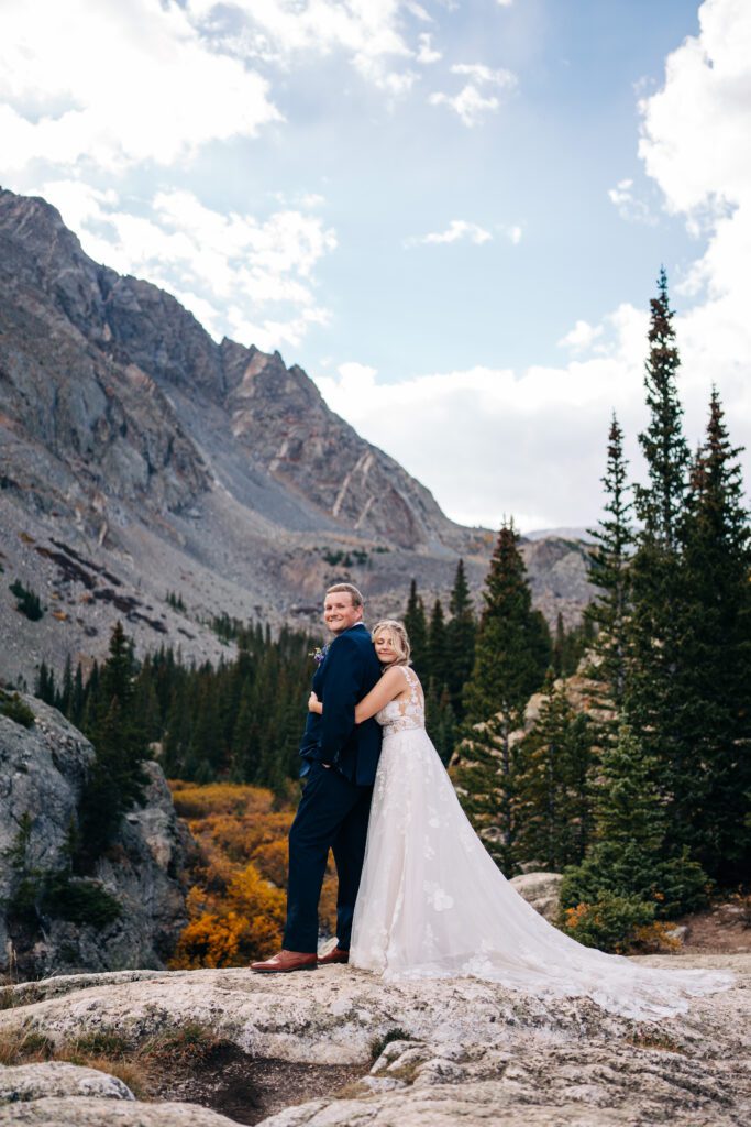 Bride hugging groom from behind as they stand on top of a rock in the Colorado mountains during their Breckenridge elopement portraits
