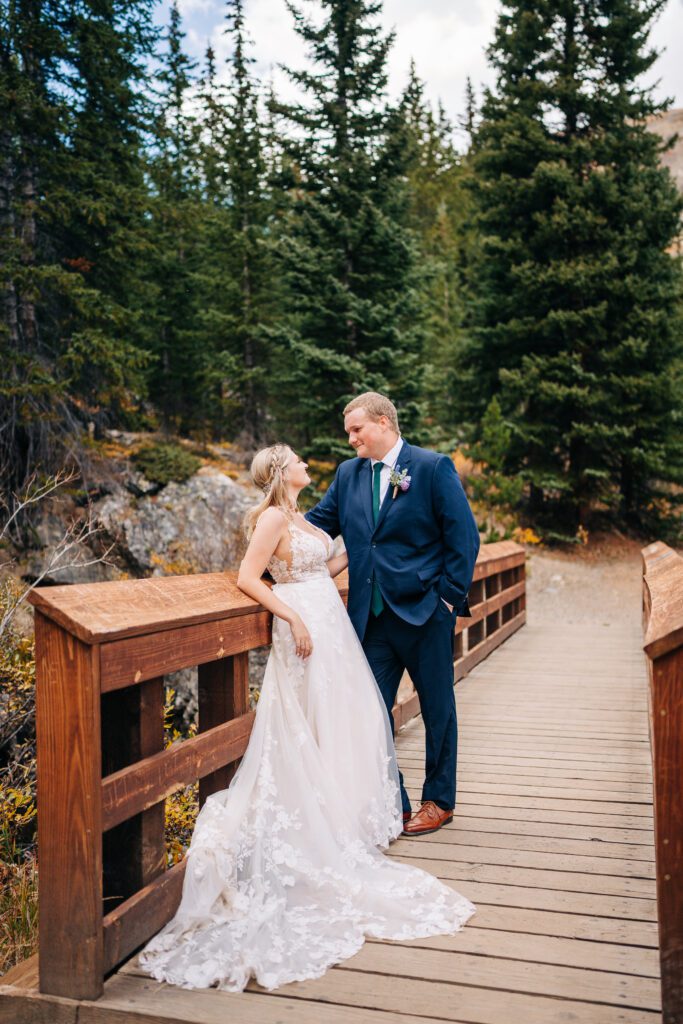 Bride leaning against the rail of a bridge as the groom smiles down at her during in a pine forest in Colorado during their Breckenridge elopement