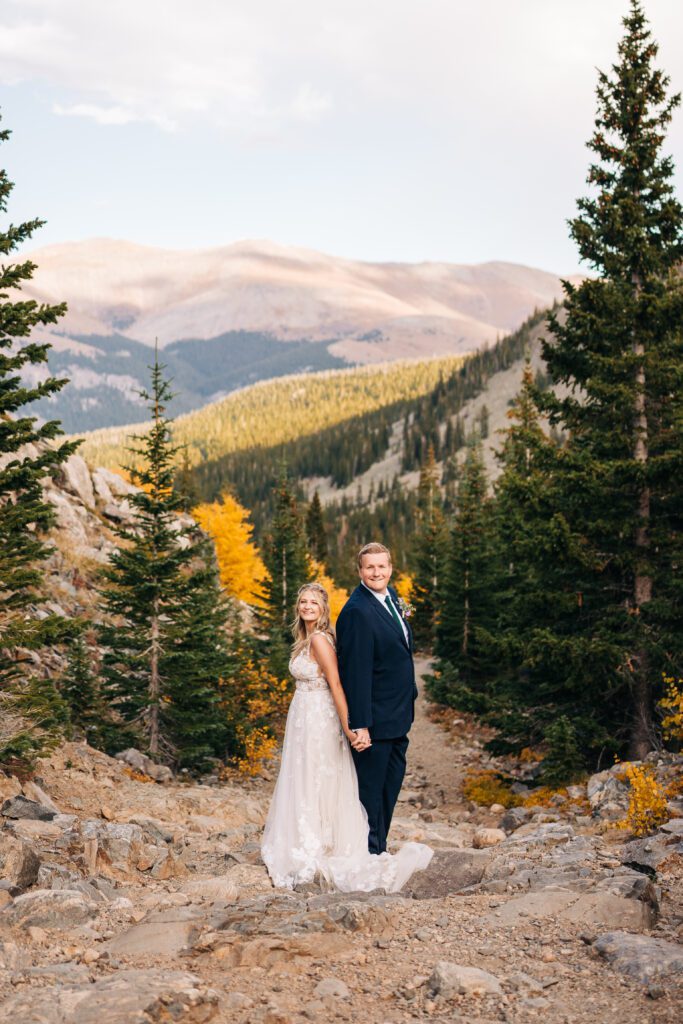 Bride and groom standing back to back smiling at the camera and holding hands in the middle of the Colorado mountains during their Breckenridge elopement