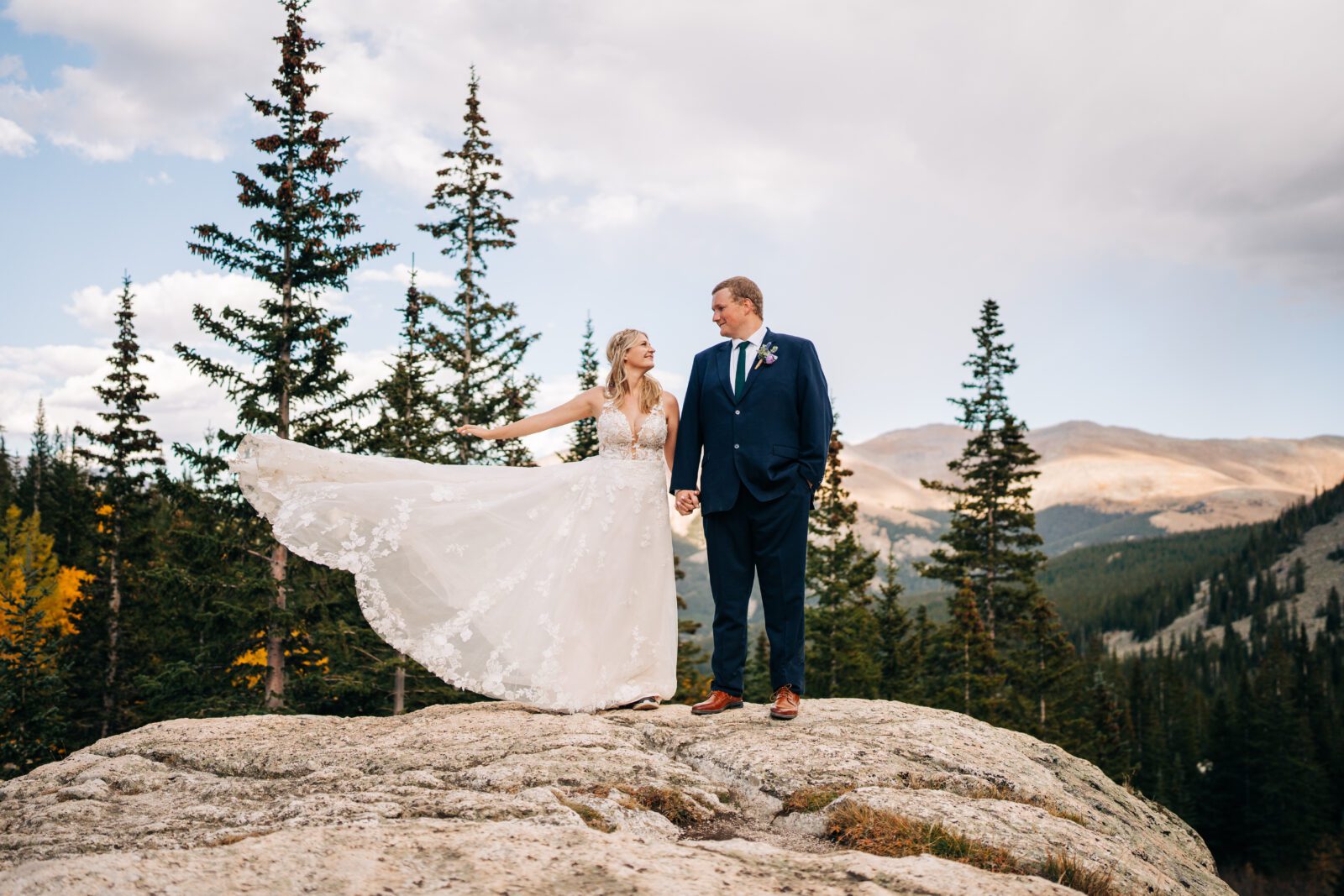 Bride throwing her dress up in the air while they stand on top of a rock in the mountains of Colorado during their bride and groom portraits after their Breckenridge elopement