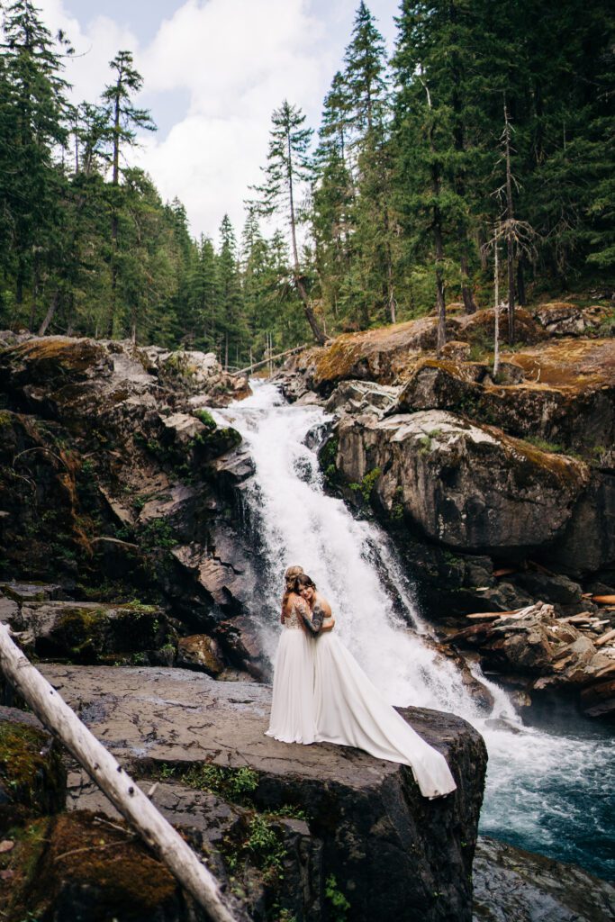 wife hugging her wife from behind on the side of cliff in front of a waterfall in the forest in mt rainier during their washington elopement 