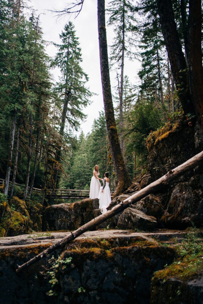 bride helping her wife up on top of a cliff after their forest wedding ceremony in   mt rainier national park during their washington elopement