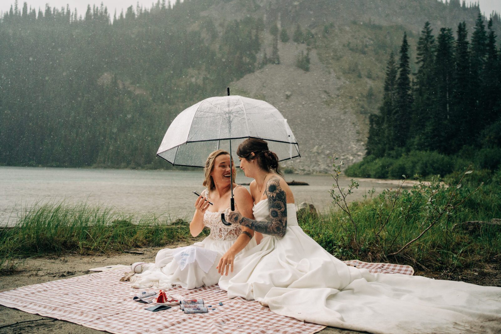 wives sitting on a blanket next to an alpine lake while it rains in mt raineir national park during their washington elopement