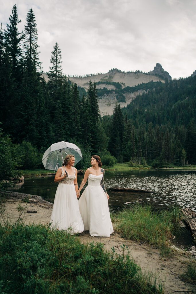 wives holding hands walking along the shore of a lake in mt rainier national park during their washington elopement