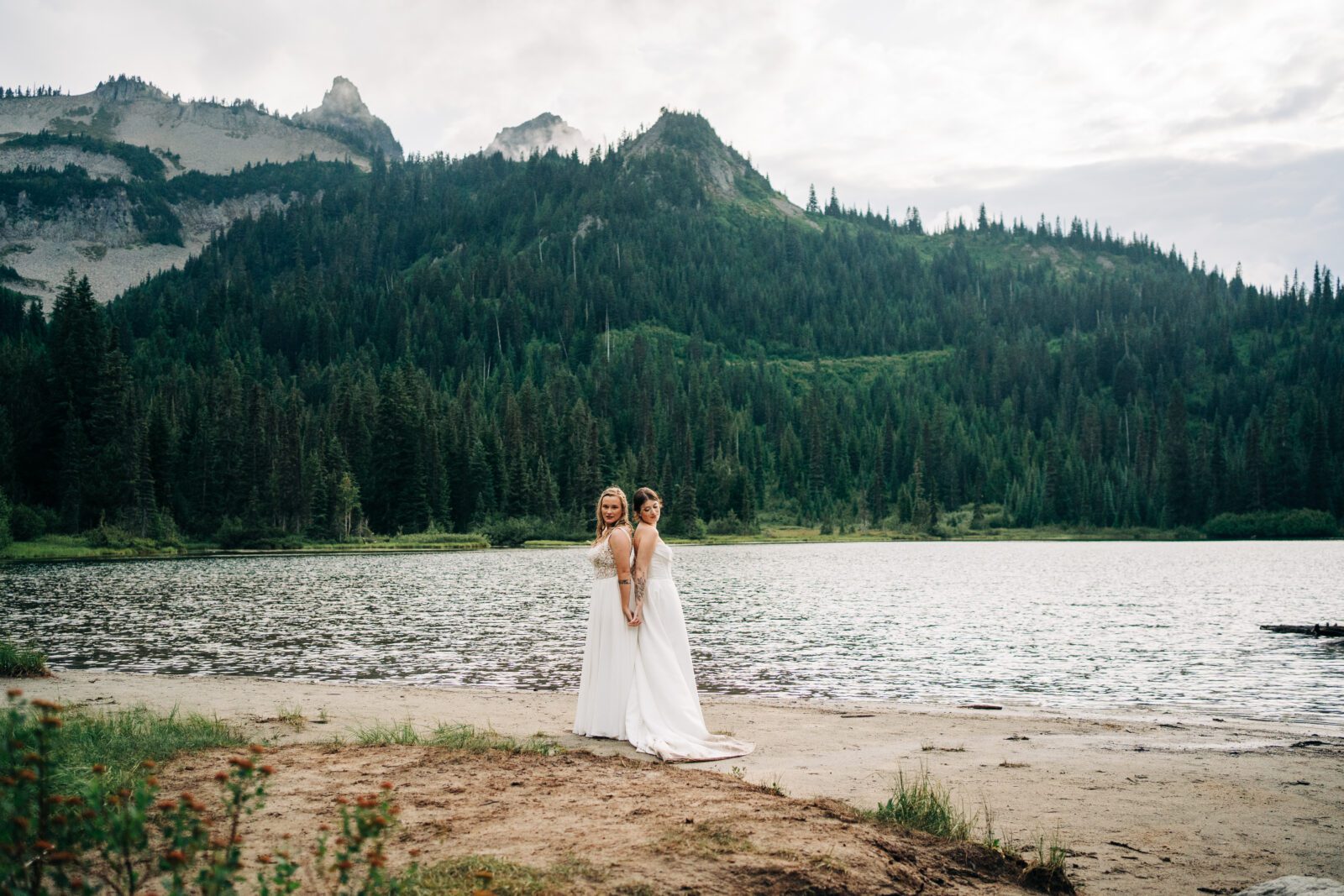 wifes standing back to back holding hands in front of an alpine lake in mt rainier national park during their washington elopement