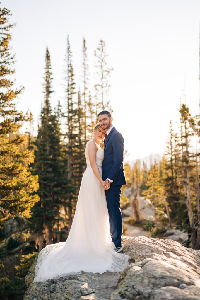 bride and groom holding hands smiling at the camera as they stand on top of a boulder during their Rocky Mountain National Park Elopement