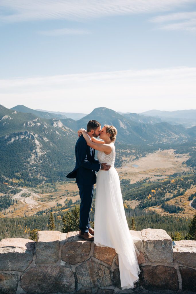 Bride and groom standing on a rock wall as they kiss overlook a valley during their colorado rocky mountain national park elopement