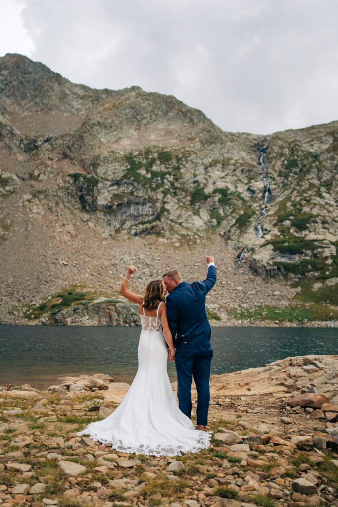 Bride and groom facing away from the camera looking at a mountain as they kiss and fist pump after their Telluride Elopement celebration at Hope Lake