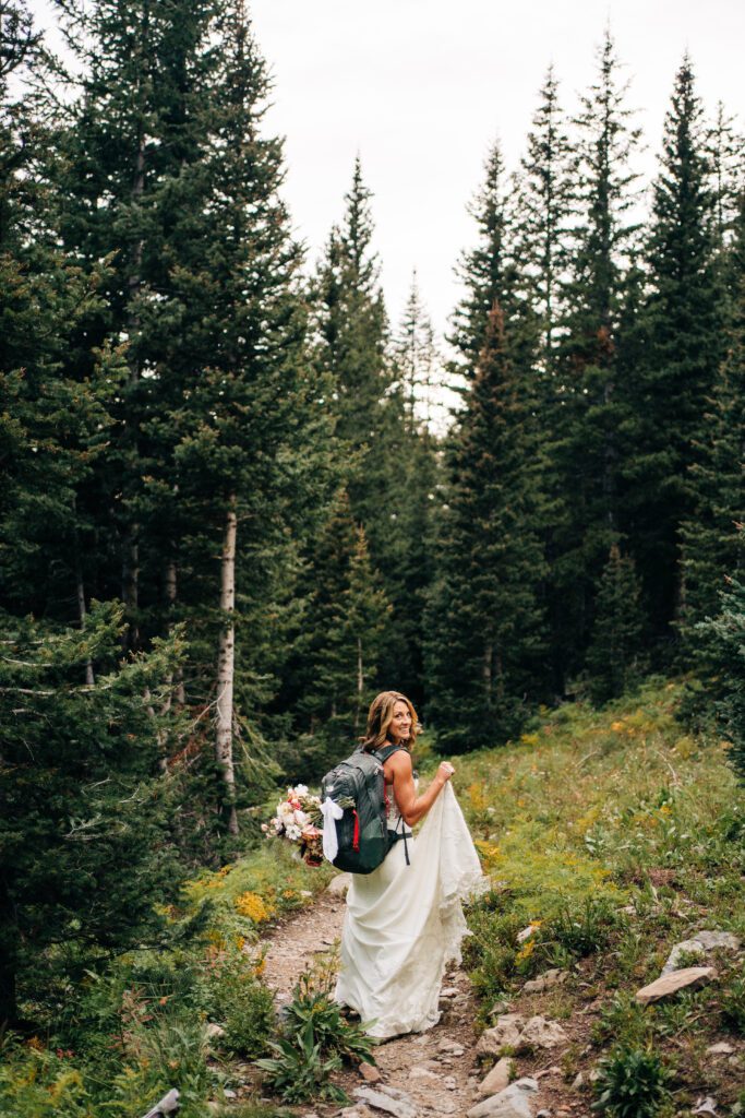 Bride with backpack and bouquet walking down a hiking trail in the san juan mountains during her Telluride elopement